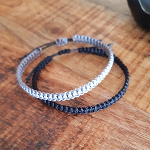 Op de afbeelding: Twee gevlochten vriendschapsarmbanden, een zwart en een grijs, liggen op een houten oppervlak.