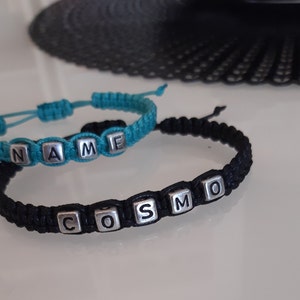 Puede incluir: Dos pulseras de la amistad con los nombres "NAME" y "COSMO" escritos en cuentas plateadas. La pulsera "NAME" es de color azul turquesa y la pulsera "COSMO" es negra.