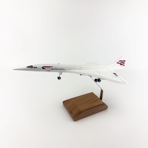 Puede incluir: Maqueta blanca de un avión Concorde de British Airways, con el logotipo en la cola y el fuselaje. El modelo está montado sobre una base de madera.