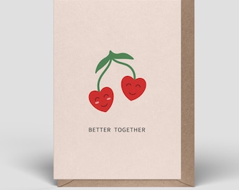 Op de afbeelding: Een lichtroze wenskaart met een bruine envelop met twee rode hartvormige kersen met groene steeltjes en blaadjes. De kersen hebben eenvoudige zwarte gezichtjes met een glimlach. De tekst BETTER TOGETHER staat in het zwart eronder gedrukt.