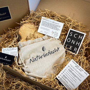 Puede incluir: Una caja de regalo llena de papel de embalaje marr&oacute;n y una tabla de cortar de madera con un dise&ntilde;o tallado. La caja tambi&eacute;n contiene una peque&ntilde;a bolsa blanca con el texto "Naturbedacht" impreso en ella y una tarjeta negra con el texto "SCH&Ouml;NHER" impreso en ella.