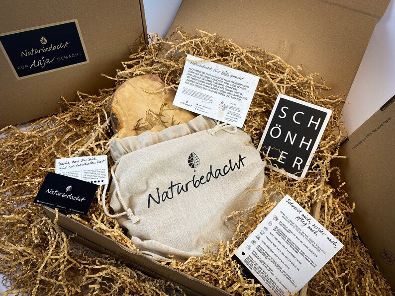 Puede incluir: Una caja de regalo llena de papel de embalaje marr&oacute;n y una tabla de cortar de madera con un dise&ntilde;o tallado. La caja tambi&eacute;n contiene una peque&ntilde;a bolsa blanca con el texto "Naturbedacht" impreso en ella y una tarjeta negra con el texto "SCH&Ouml;NHER" impreso en ella.