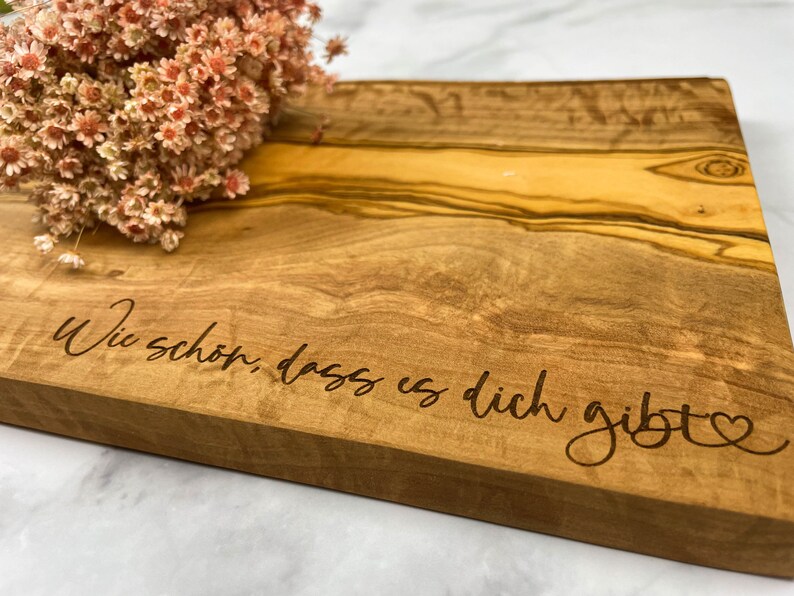 Puede incluir: Una tabla de cortar de madera con un acabado de grano de madera natural. La tabla tiene un coraz&oacute;n y el texto "Wie sch&ouml;n, dass es dich gibt" grabado en la superficie.