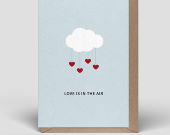 Op de afbeelding: Een lichtblauwe wenskaart met een witte wolk-afbeelding waaraan vier kleine rode hartjes hangen aan dunne lijntjes. Onder de wolk staat de tekst LOVE IS IN THE AIR in het zwart gedrukt. De kaart ligt op een bruine envelop.