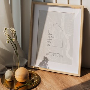 May include: A framed print showing a map of D&uuml;sseldorf, Germany, with a house outline and names. The print is light grey, with the city name and coordinates at the bottom. The frame is light wood.