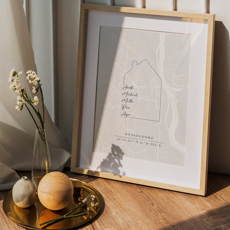 May include: A framed print showing a map of D&uuml;sseldorf, Germany, with a house outline and names. The print is light grey, with the city name and coordinates at the bottom. The frame is light wood.