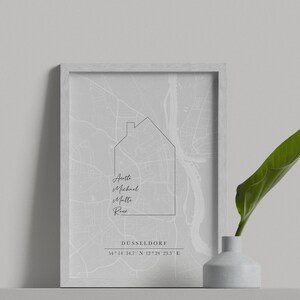 May include: A framed print showing a grey map of D&uuml;sseldorf, Germany, with a house outline. Names are below the house graphic, with the city name and coordinates at the bottom. A white vase with a green leaf is on the right.