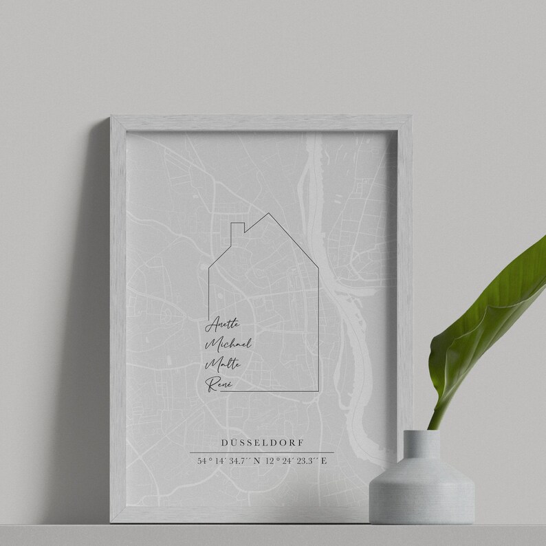 May include: A framed print showing a grey map of D&uuml;sseldorf, Germany, with a house outline. Names are below the house graphic, with the city name and coordinates at the bottom. A white vase with a green leaf is on the right.