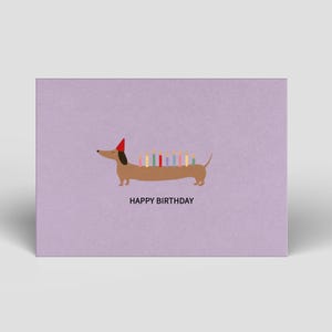 Peut inclure: Carte d'anniversaire sur fond lilas. Un teckel marron avec un chapeau de fête rouge porte des bougies colorées sur le dos. L'inscription "HAPPY BIRTHDAY" est imprimée en noir sous le chien.