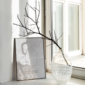 May include: A framed gray map print with the text "Holle More" and "Frankfurt am Main" is displayed. A decorative branch with thorns is in a clear glass vase with a white dotted pattern. The items are on a windowsill.