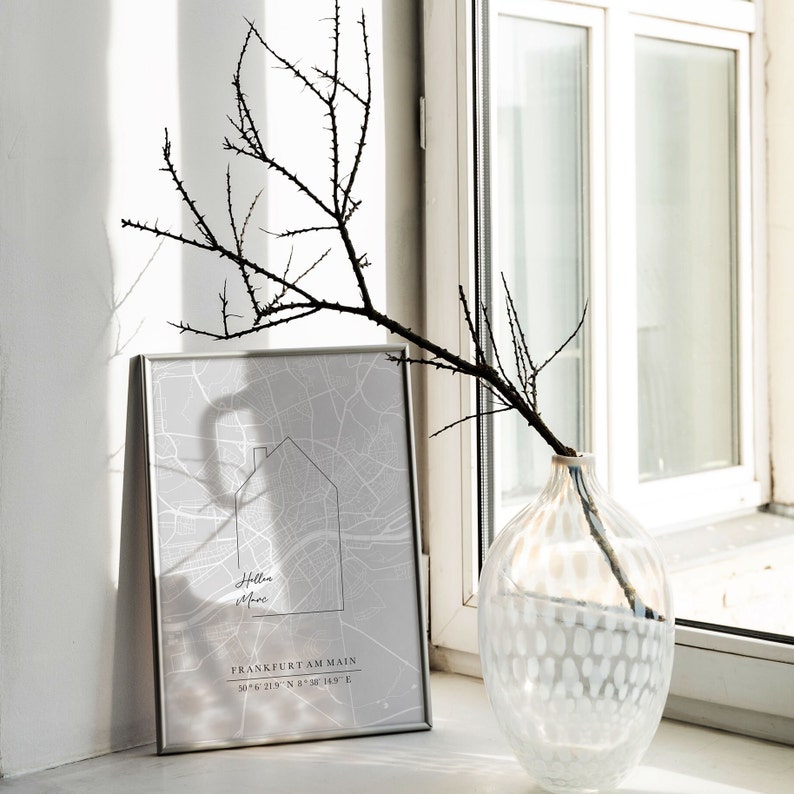 May include: A framed gray map print with the text "Holle More" and "Frankfurt am Main" is displayed. A decorative branch with thorns is in a clear glass vase with a white dotted pattern. The items are on a windowsill.