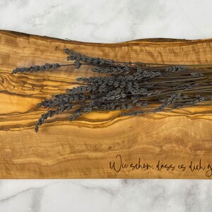 Puede incluir: Una tabla de servir de madera con un patr&oacute;n de veta de madera natural. La tabla tiene una forma ligeramente curvada y est&aacute; grabada con el texto "Wie sch&ouml;n, dass es dich gibt" en una fuente cursiva. Un manojo de lavanda seca se coloca encima de la tabla.