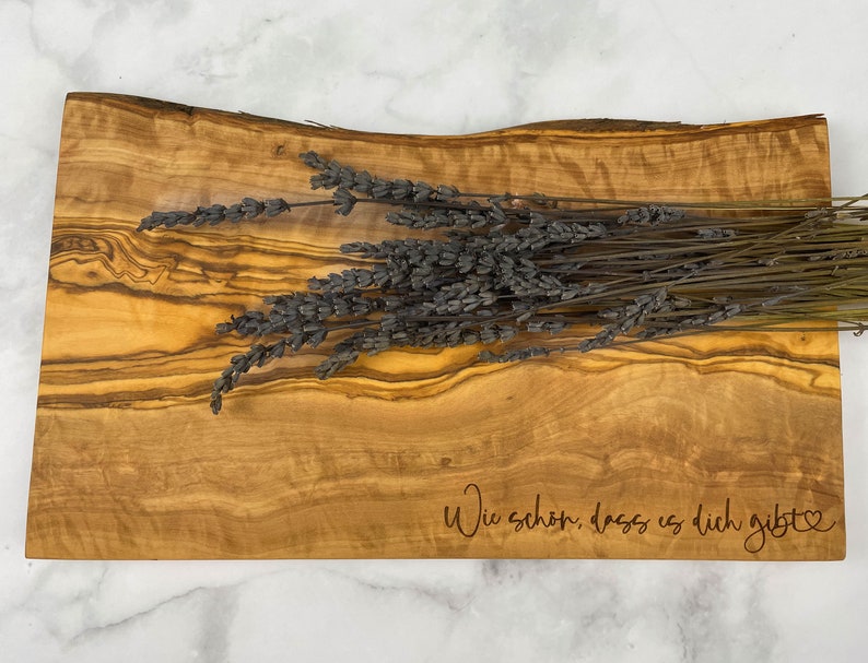 Puede incluir: Una tabla de servir de madera con un patr&oacute;n de veta de madera natural. La tabla tiene una forma ligeramente curvada y est&aacute; grabada con el texto "Wie sch&ouml;n, dass es dich gibt" en una fuente cursiva. Un manojo de lavanda seca se coloca encima de la tabla.