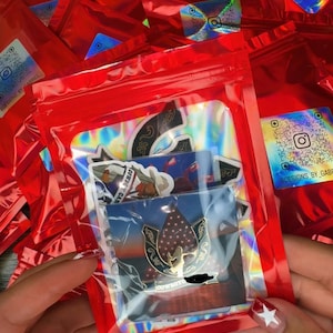 May include: A clear plastic bag containing stickers and pins, held in front of a pile of red plastic bags. The stickers and pins have various designs, including a cowboy hat and a horseshoe. The bags have a holographic sticker with a social media handle.