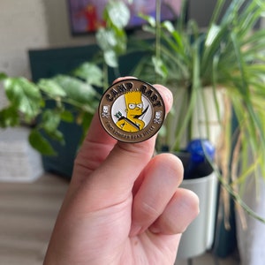 May include: A gold-colored pin with a cartoon image of a yellow-haired character holding a bow and arrow. The pin says "Camp Bart" and "I no longer fear heights" in black text.