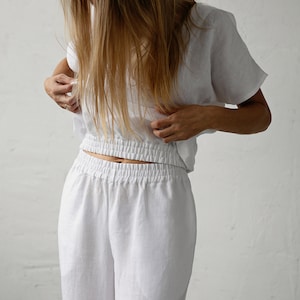May include: White linen two-piece set. The top is a short-sleeved, cropped style with a gathered waist. The pants are wide-legged with an elastic waistband. The fabric appears lightweight and breathable, suitable for warm weather.