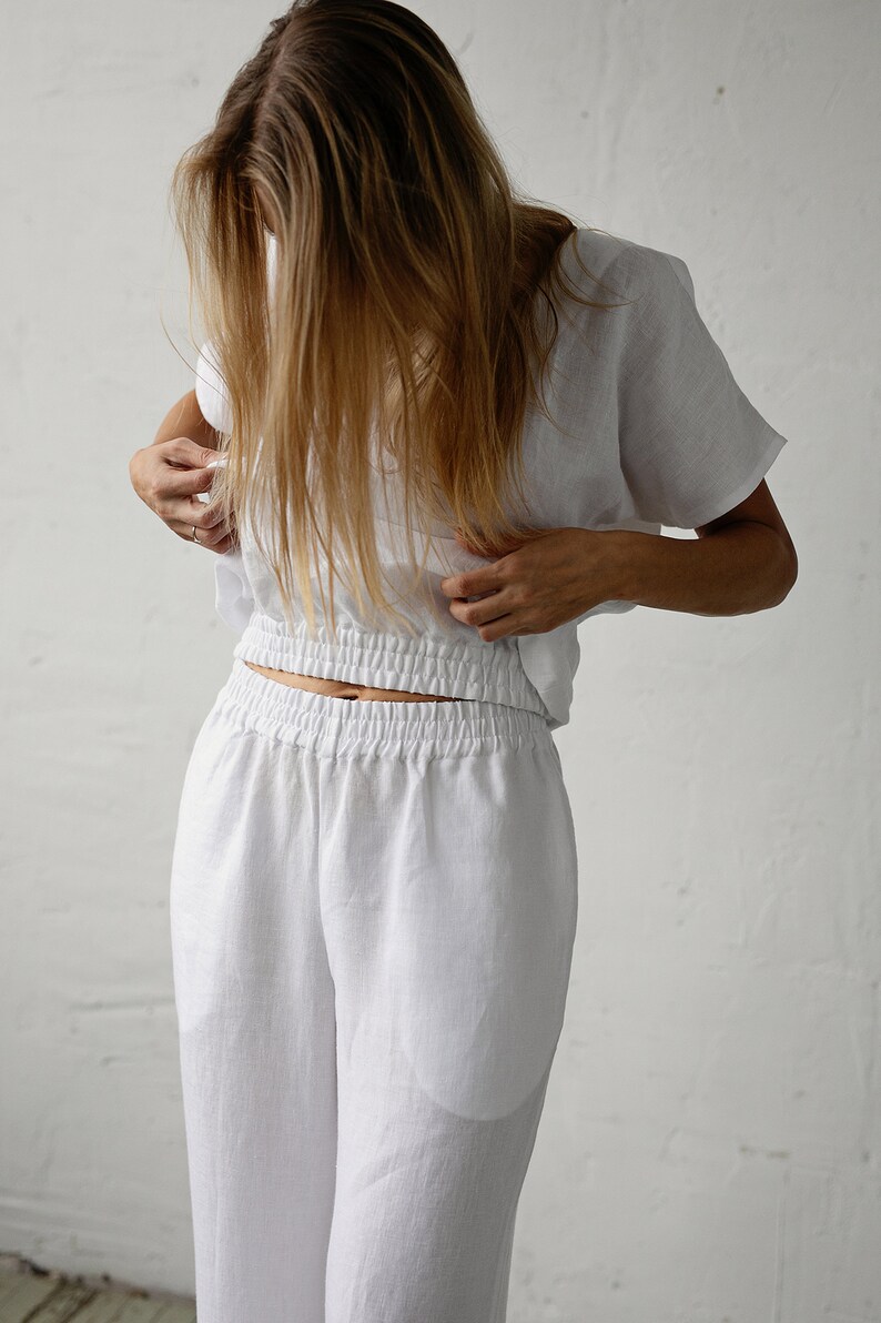 May include: White linen two-piece set. The top is a short-sleeved, cropped style with a gathered waist. The pants are wide-legged with an elastic waistband. The fabric appears lightweight and breathable, suitable for warm weather.