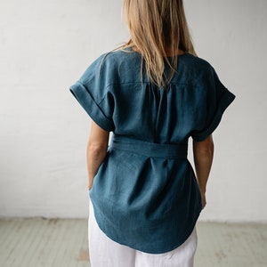 May include: A teal linen top with a tie belt and short sleeves. The top is loose fitting and has a gathered detail at the back.