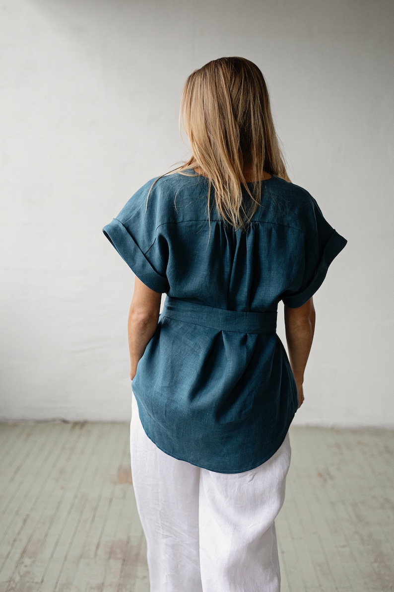 May include: A teal linen top with a tie belt and short sleeves. The top is loose fitting and has a gathered detail at the back.