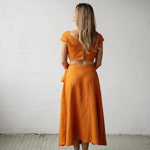 May include: A two-piece outfit in a burnt orange colour. The top is a cropped top with short sleeves and a V-neckline. The skirt is a midi-length skirt with a wrap style and a flowing silhouette.