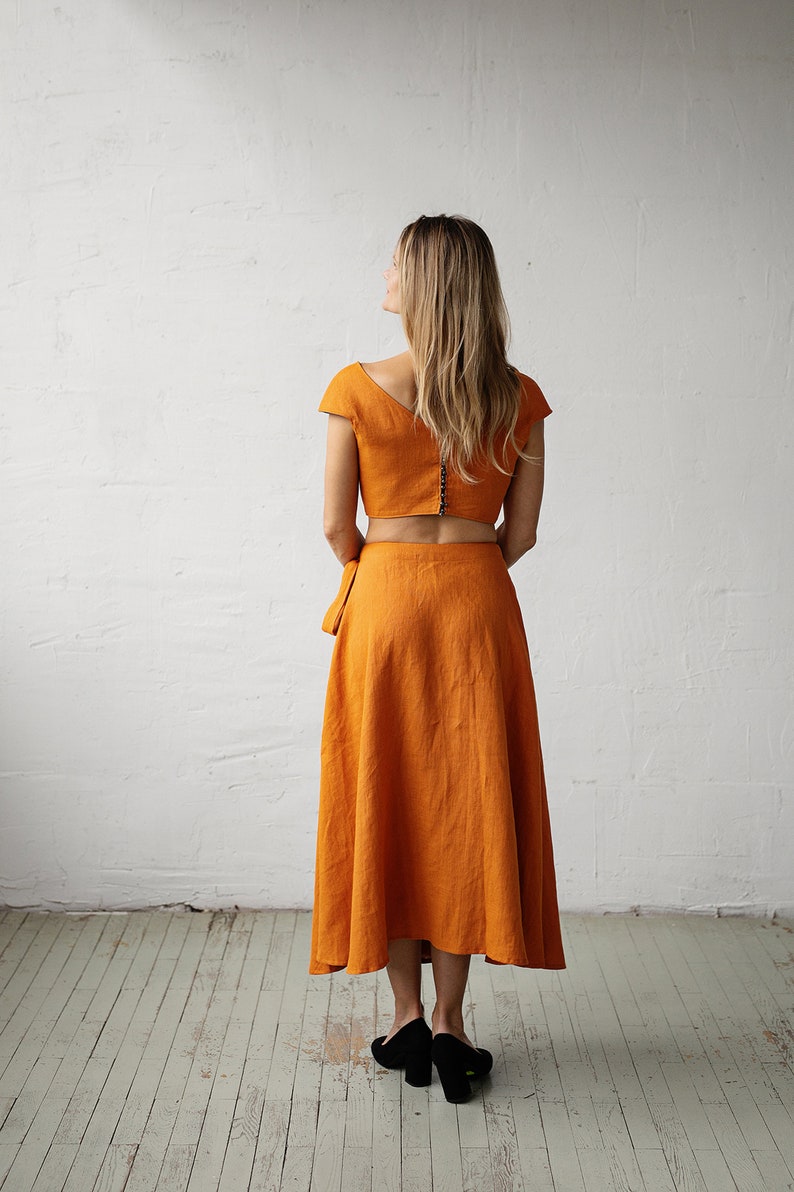 May include: A two-piece outfit in a burnt orange colour. The top is a cropped top with short sleeves and a V-neckline. The skirt is a midi-length skirt with a wrap style and a flowing silhouette.