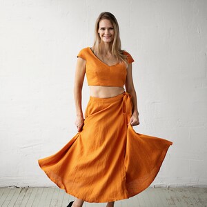 May include: A woman wearing a two-piece outfit in a burnt orange colour. The top is a cropped, fitted style with a V-neckline and short sleeves. The skirt is a wrap style with a flowing silhouette. She is wearing black heels.