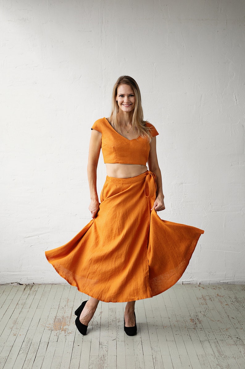 May include: A woman wearing a two-piece outfit in a burnt orange colour. The top is a cropped, fitted style with a V-neckline and short sleeves. The skirt is a wrap style with a flowing silhouette. She is wearing black heels.