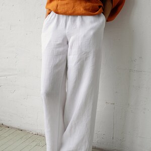 May include: White linen pants with a wide leg design. The pants are paired with an orange top. The model is wearing beige sandals. The outfit is set against a white wall and a light green wooden floor.