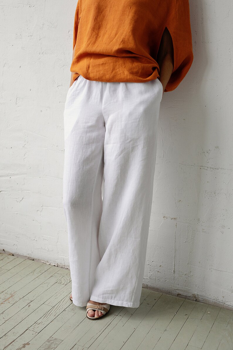 May include: White linen pants with a wide leg design. The pants are paired with an orange top. The model is wearing beige sandals. The outfit is set against a white wall and a light green wooden floor.