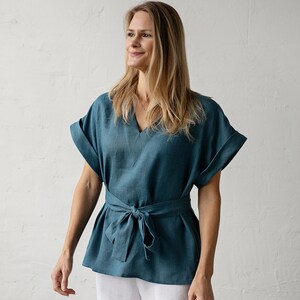 May include: A teal linen top with a V-neck and short sleeves. The top has a tie at the waist and is worn with white linen pants.