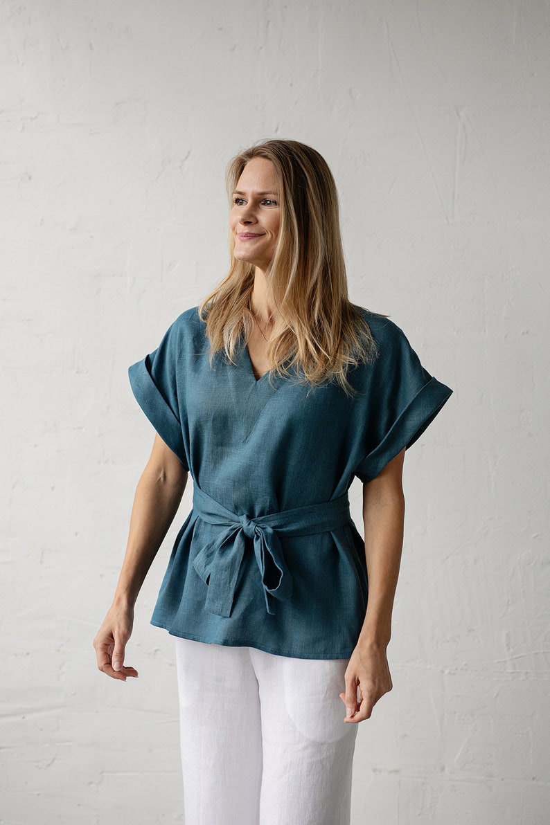May include: A teal linen top with a V-neck and short sleeves. The top has a tie at the waist and is worn with white linen pants.