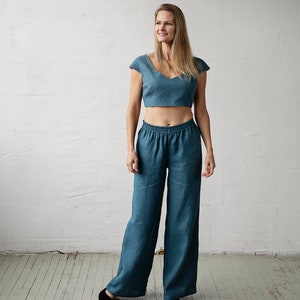 May include: A teal two-piece linen outfit. The top is a cropped, cap-sleeved design with a V-neck. The matching pants are wide-legged with an elastic waistband. The outfit is worn with black flats.