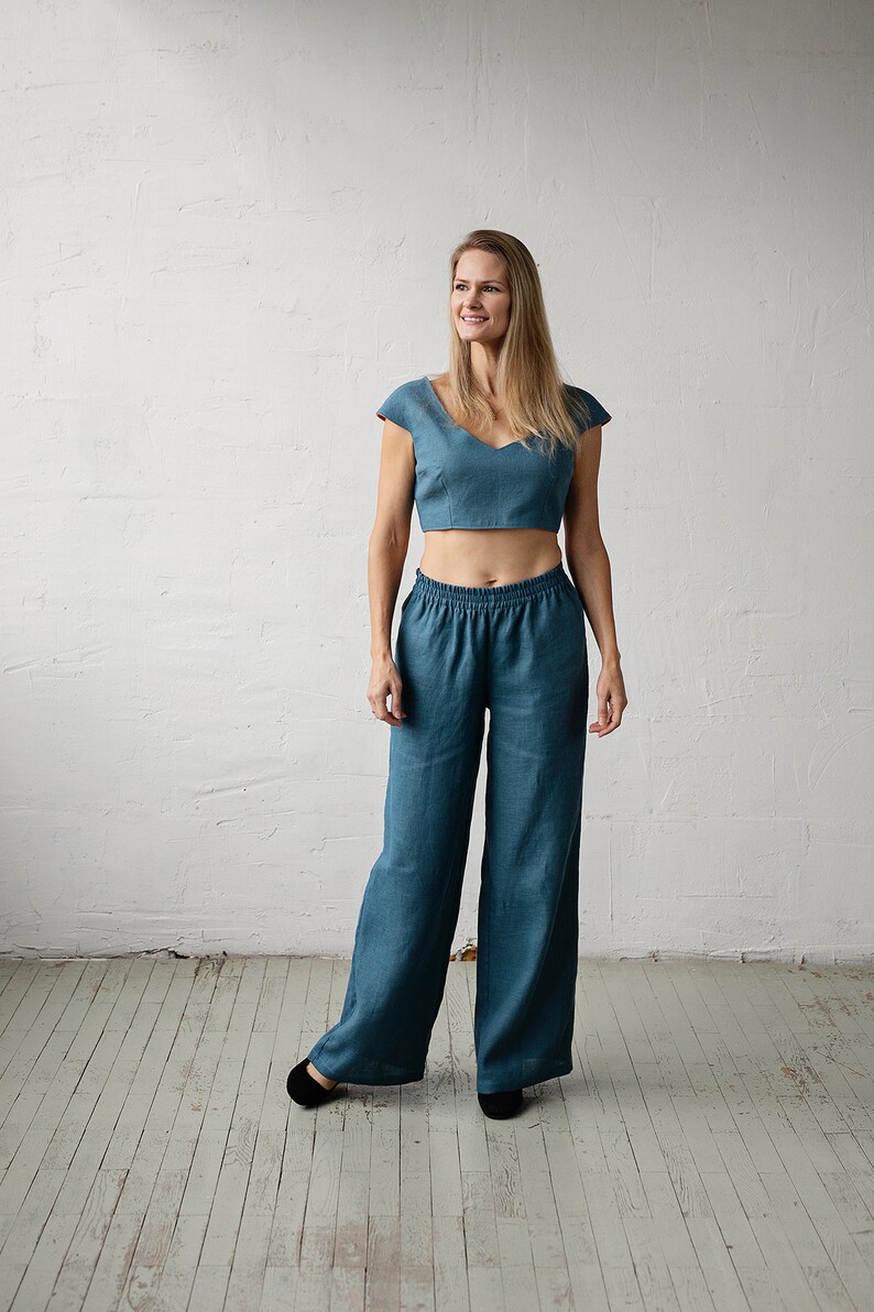May include: A teal two-piece linen outfit. The top is a cropped, cap-sleeved design with a V-neck. The matching pants are wide-legged with an elastic waistband. The outfit is worn with black flats.