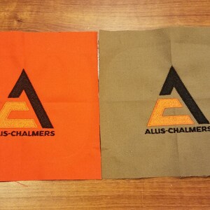 Puede incluir: Dos parches de tela con el logotipo de Allis-Chalmers. El logotipo es una A estilizada con una E más pequeña en su interior. El parche de la izquierda es naranja y el parche de la derecha es beige.