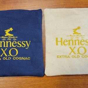 May include: Two square fabric pouches, one navy blue and one off-white, each featuring yellow embroidered text that reads Maison Fondée en 1765, Hennessy X.O, and EXTRA OLD COGNAC, with a small graphic icon above the brand name.