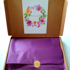 May include: A brown cardboard box with a purple tissue paper lining. The box is open and a white card with a watercolor floral wreath is visible. The card says "Bloom where you're planted". A small round sticker on the tissue paper says "handmade with love".