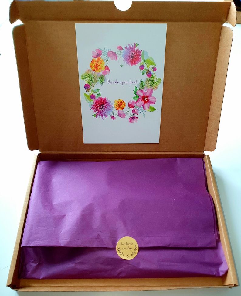 May include: A brown cardboard box with a purple tissue paper lining. The box is open and a white card with a watercolor floral wreath is visible. The card says "Bloom where you're planted". A small round sticker on the tissue paper says "handmade with love".