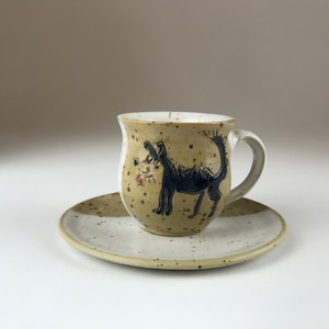 Op de afbeelding: Een keramische koffiekop en schotelset met een gespikkeld bruin en wit glazuur. De kop heeft een handgeschilderde illustratie van een zwarte hond met een rode neus.