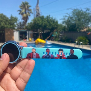 May include: A blue smartwatch with a black screen and a band featuring a print of characters from the Marvel Cinematic Universe. The watch is being held by a hand in front of a swimming pool.