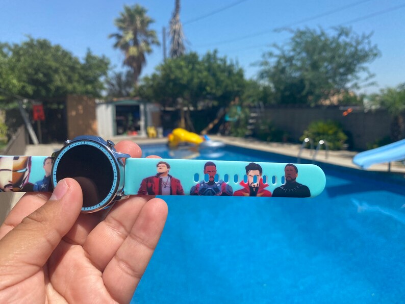 May include: A blue smartwatch with a black screen and a band featuring a print of characters from the Marvel Cinematic Universe. The watch is being held by a hand in front of a swimming pool.