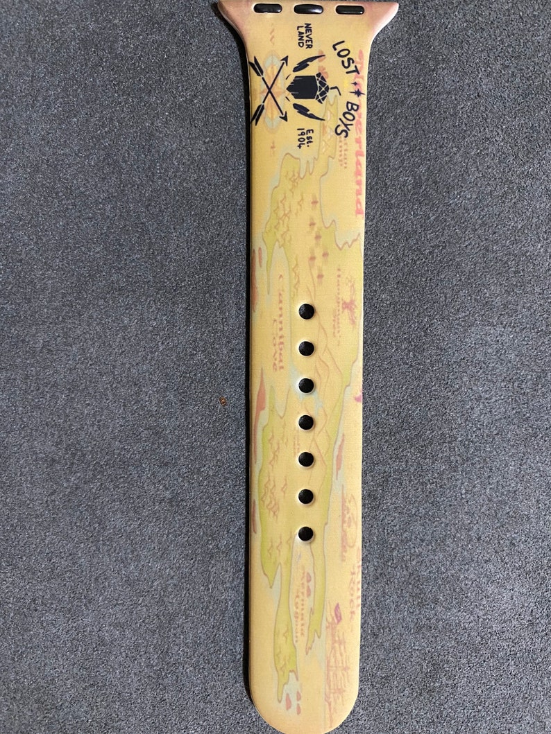 Puede incluir: Correa de reloj Apple Watch de color beige con un dise&ntilde;o de mapa y las palabras "Lost Boys" y "Never Land". La correa incluye una br&uacute;jula y flechas cruzadas, as&iacute; como el texto "Est. 1904."