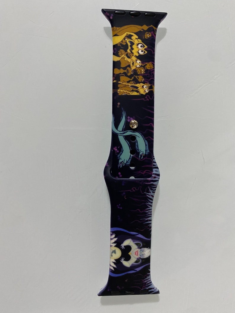 May include: A black and purple Apple Watch band with a graphic print of characters from the Disney movie The Little Mermaid. The band features Ursula, the sea witch, and the sea creatures from the movie.