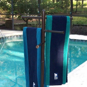 May include: A brown metal towel rack with two towels hanging on it. The towels are blue and green with white text that says "Ocean Lamp". The rack is standing next to a swimming pool.