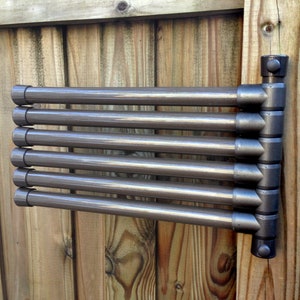 May include: A brown metal drying rack with six horizontal bars attached to a vertical support. The rack is mounted to a wooden fence.