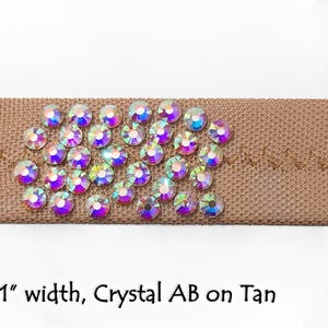 May include: A close-up of a 1 inch wide tan ribbon with crystal AB rhinestones. The rhinestones are arranged in a cluster and have a rainbow iridescent effect.