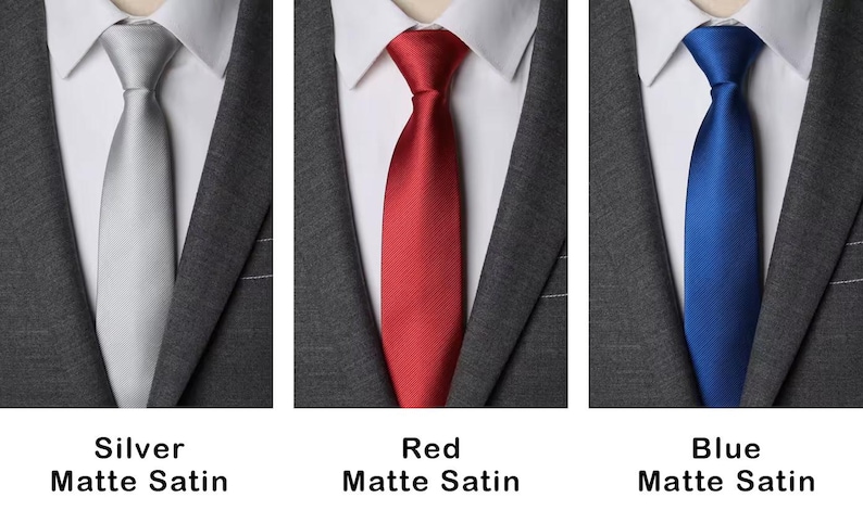 K&ouml;nnte beinhalten: Drei Krawatten in Silber, Rot und Blau, jeweils mit matter Satin-Oberfl&auml;che. Die Krawatten sind gebunden und werden &uuml;ber einem wei&szlig;en Hemd mit Kragen und einem dunkelgrauen Sakko getragen. Text unter jeder Krawatte benennt die Farbe.