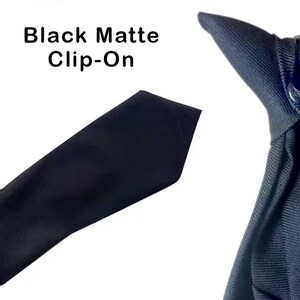 K&ouml;nnte beinhalten: Eine schwarze, matte Clip-Krawatte aus einfarbigem, dunklem Stoff mit strukturierter Oberfl&auml;che. Der obere Teil enth&auml;lt einen funktionalen schwarzen Kunststoff-Clip zur Befestigung. Der Text Black Matte Clip-On ist im Bild zu sehen.