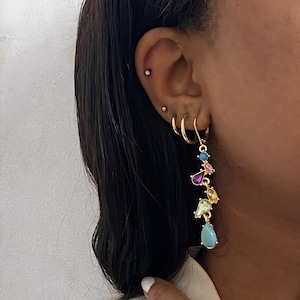 Pode incluir: Brincos de argola dourados com um brinco pendente que apresenta uma cascata de pedras preciosas coloridas em forma de lágrima. O brinco pendente apresenta uma mistura de pedras preciosas azuis, verdes, roxas e amarelas.