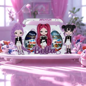 Puede incluir: Una caja blanca decorativa con tapa rosa, con figuras de dibujos animados y huevos Kinder Sorpresa. El nombre "Bella" está escrito en letras moradas. El fondo es rosa suave con elementos decorativos.