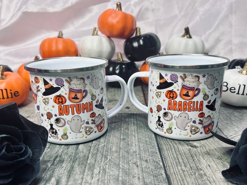 K&ouml;nnte beinhalten: Zwei wei&szlig;e Emailletassen mit Halloween-Motiven. Eine Tasse sagt "Autumn" und die andere sagt "Arabella".